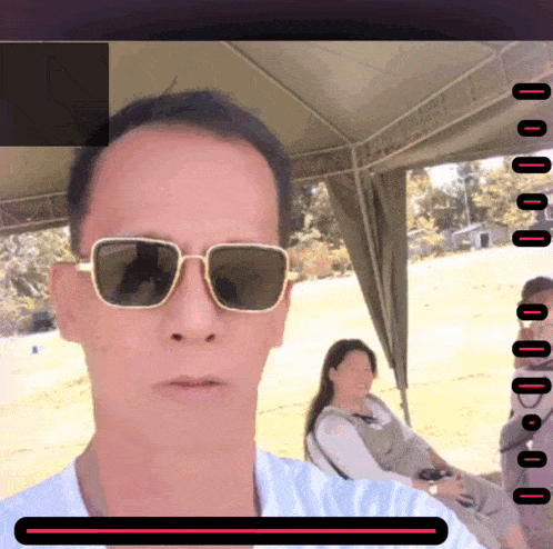 a man wearing sunglasses takes a selfie with a woman sitting in the background