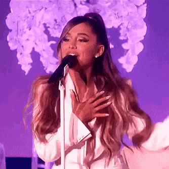 ariana grande is singing into a microphone on stage while wearing a white suit .