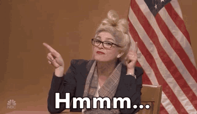 a woman in a wig and glasses is sitting in front of an american flag .