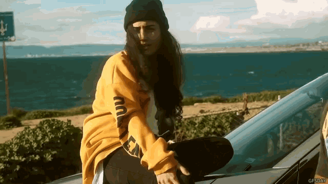 a woman wearing a yellow sweatshirt that says ea on the sleeve sits on the hood of a car
