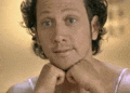 a man with curly hair and a beard is holding his hands to his face .