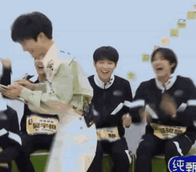 a group of young men are sitting on a green bench and laughing while a man stands in front of them .