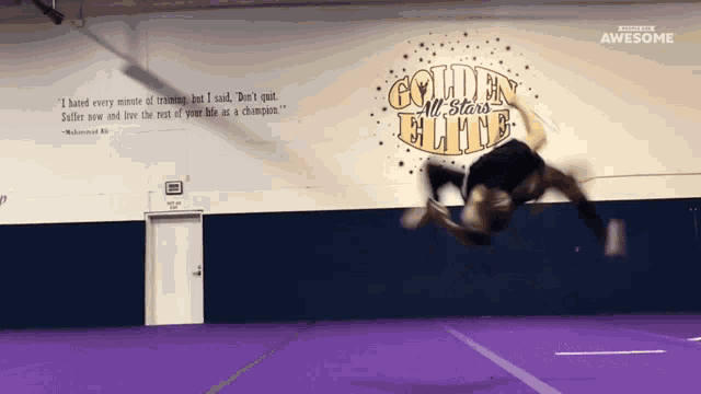 a cheerleader is doing a trick in front of a wall that says golden all stars elite