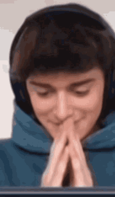 a man wearing headphones and a blue hoodie is praying with his hands folded in front of a computer screen .