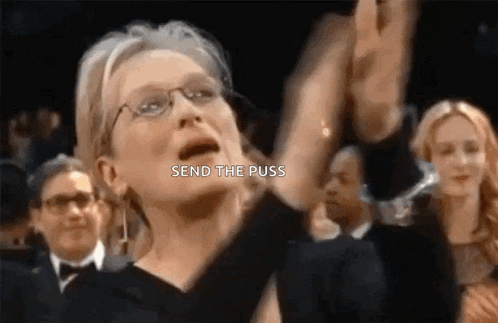 a woman wearing glasses is sitting in a crowd with her hands in the air and screaming .