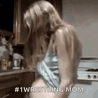 a woman is standing in a kitchen taking off her shirt and says `` # 1 wrestling mom '' .