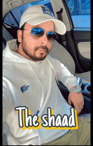 a man wearing a hat and sunglasses is sitting in a car with the shaad written on the bottom right