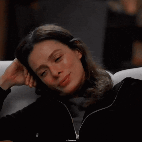 a woman in a black jacket smiles while sitting on a white couch with her eyes closed