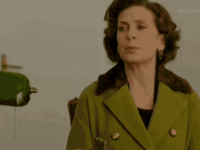 a woman in a green coat stands in front of a green lamp with the letters agriasm on the bottom