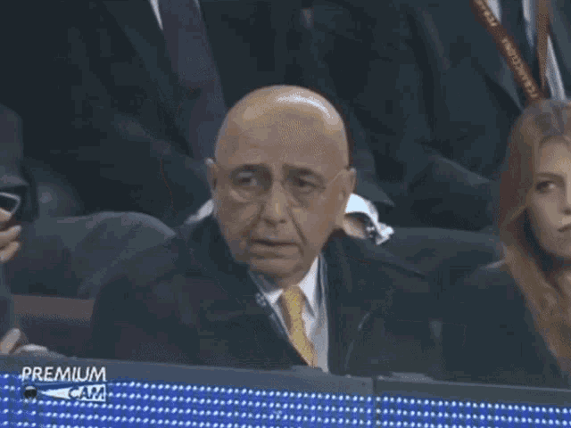 a man in a suit and tie sits in a stadium with a sign that says futbol karimm on it
