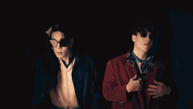 a man wearing sunglasses stands next to another man in a suit