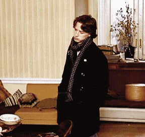 a man in a black coat and scarf stands in front of a couch