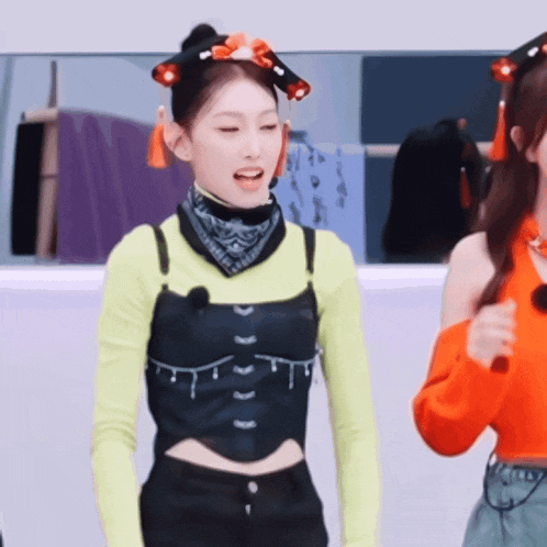 a woman wearing a yellow top and a black vest stands next to another woman in an orange top