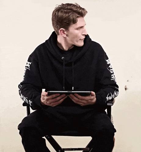 a man wearing a black hoodie with chinese writing on the sleeves is sitting on a chair holding a tablet