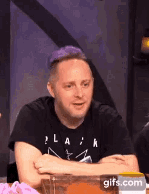 a man with purple hair is sitting at a table with his arms crossed and looking at the camera .
