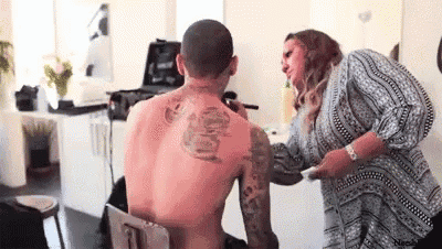 a man without a shirt is getting his back tattooed by a woman .