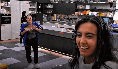 two women are standing in a kitchen and one is wearing a blue jacket that says adidas on it
