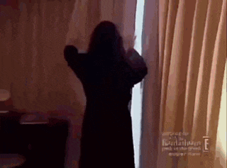 a woman in a black dress is standing in front of a window with curtains .