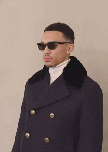 a man wearing sunglasses and a black coat has a white turtleneck underneath