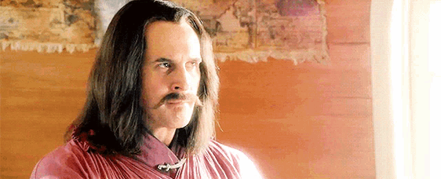 a man with long hair and a mustache is wearing a pink shirt and standing in front of a window .