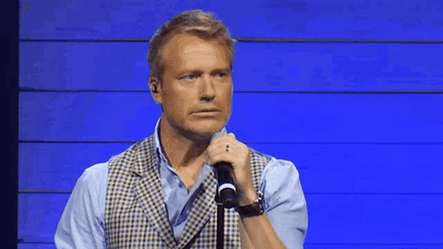 a man in a vest is holding a microphone in front of a blue wall