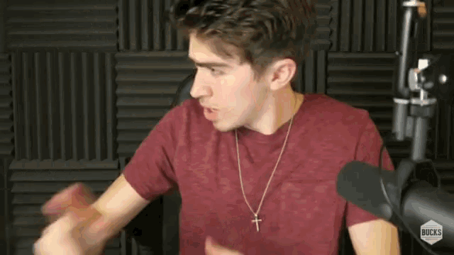a young man wearing a red shirt and a cross necklace is standing in front of a microphone .