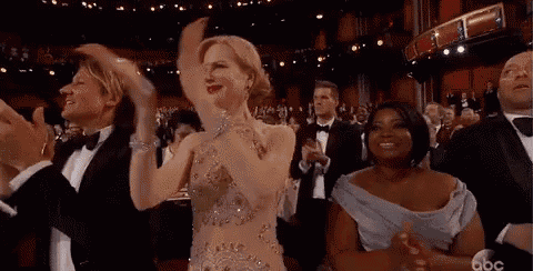a woman in a dress is applauding while standing in front of a crowd of people in tuxedos .