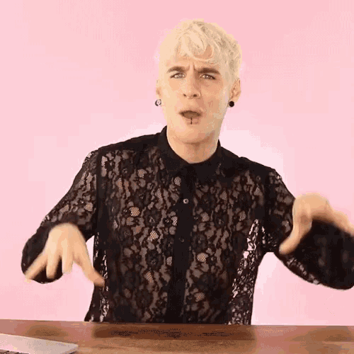 a man in a black lace shirt is sitting at a table with his hands outstretched .