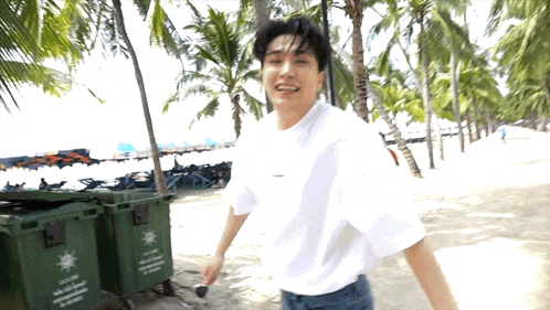 a man in a white shirt is walking on a beach next to a green trash can that says ' recycle ' on it