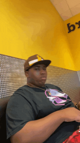 a man wearing a hat and a black shirt is sitting in front of a yellow wall with a letter b on it