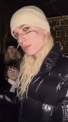 a woman with long blonde hair wearing a white beanie and a black jacket is standing in front of a group of people .