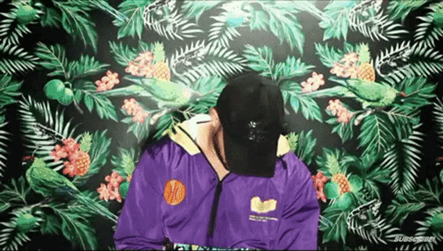 a man wearing a purple jacket and a black hat is standing in front of a tropical wall .