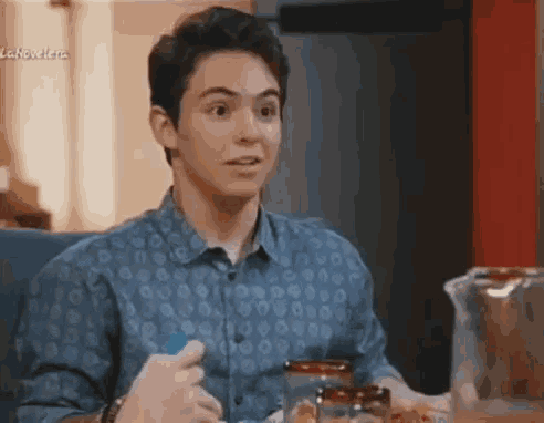a young man in a blue shirt is sitting at a table with a pitcher of water and a jar of jam .