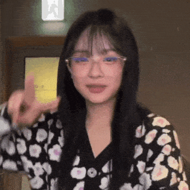 a young woman wearing glasses and a floral sweater is giving a peace sign .