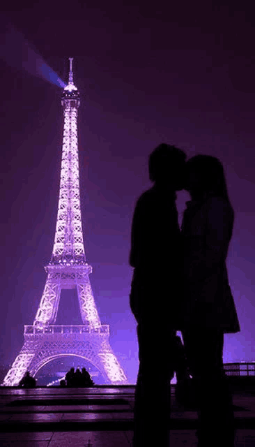 a man and a woman kissing in front of the eiffel tower at night .