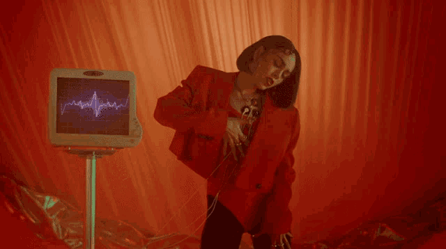 a woman in a red jacket is standing in front of a monitor that shows a heartbeat on it