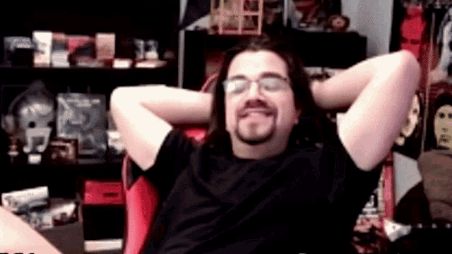 a man with long hair and glasses is sitting in a chair with his hands behind his head and smiling .