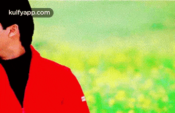 a man in a red jacket and black turtleneck is standing in a field of yellow flowers .
