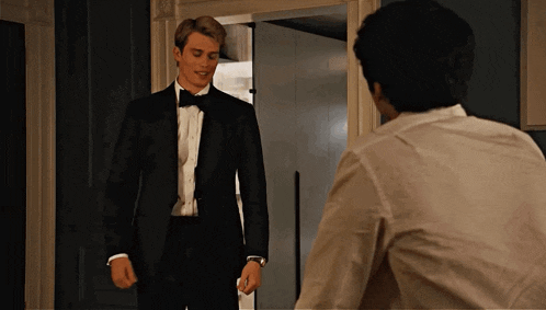a man in a tuxedo and bow tie stands next to another man in a white shirt