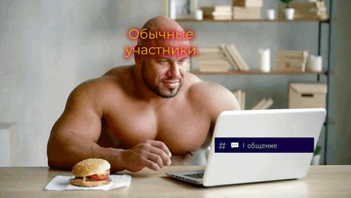 a shirtless man sits at a table with a laptop and a hamburger on a napkin in front of him
