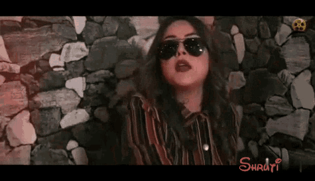 a woman wearing sunglasses and a striped shirt stands in front of a stone wall .
