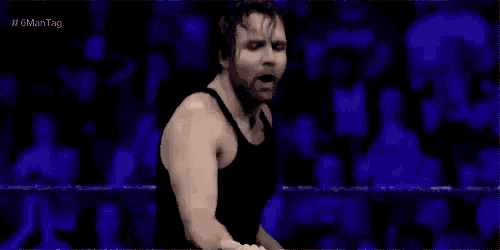 a wrestler in a black tank top is standing in a wrestling ring in front of a crowd .