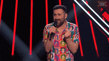 a man in a colorful shirt singing into a microphone with the x factor logo in the background