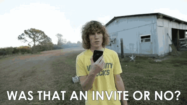 a man wearing a yellow shirt that says varsity sucks holds a cell phone in front of a barn