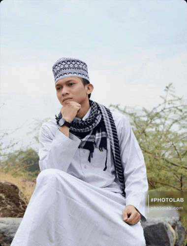 a man wearing a white robe and a black and white hat is sitting on a rock