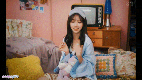 a girl is sitting on a couch with a teddy bear and a tv in the background with the letters s on the bottom