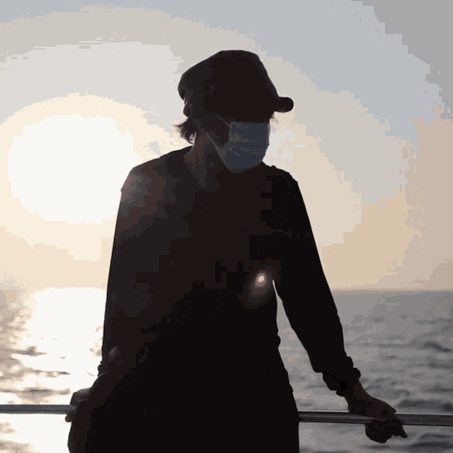 a person wearing a face mask and a hat stands in front of the ocean