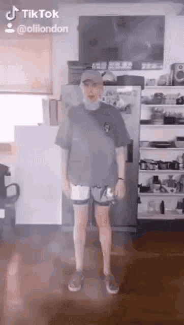 a man in a gray shirt and shorts is standing in a kitchen .