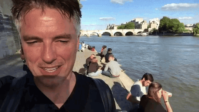 a man smiles in front of a group of people sitting on the shore of a river