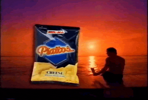 a man sits on the beach next to a bag of piatto 's cheese potato chips